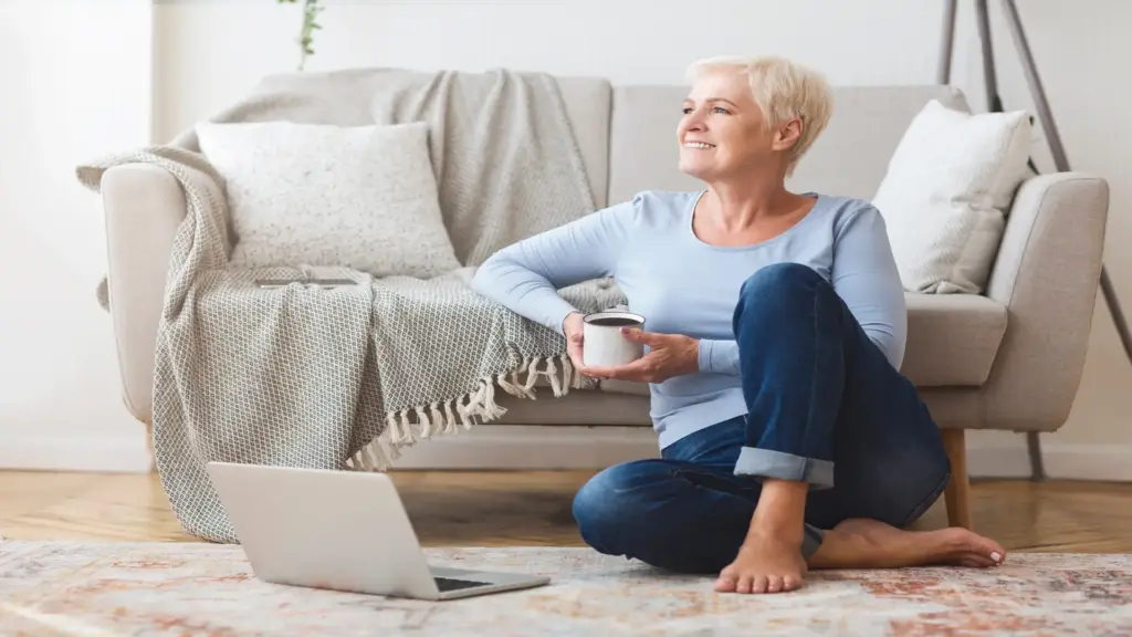 Mid-age woman sitting on the floor near a sofa, smiling while enjoying a cup of coffee.